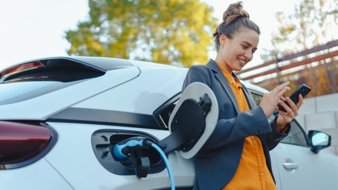 Young woman with smartphone waiting while her electric car charging in home charging station, sustainable and economic transportation concept.