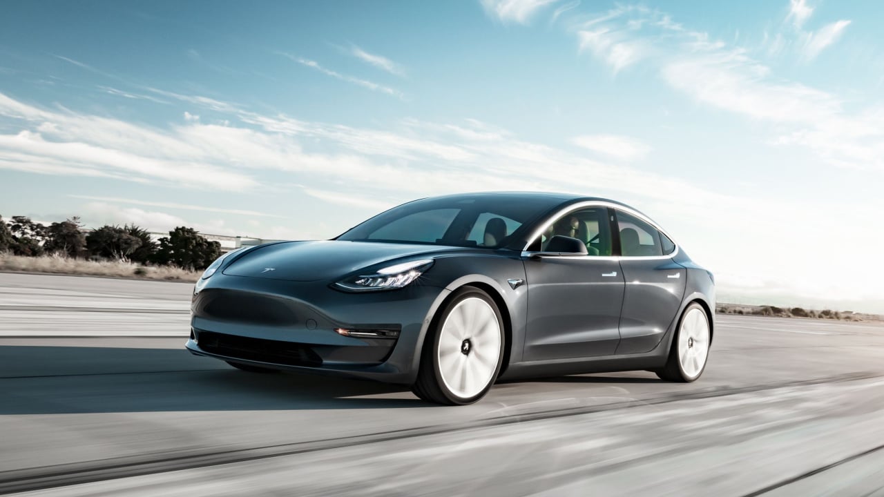 Photograph of a grey Tesla model 3 driving on the road with the sun setting in the background.