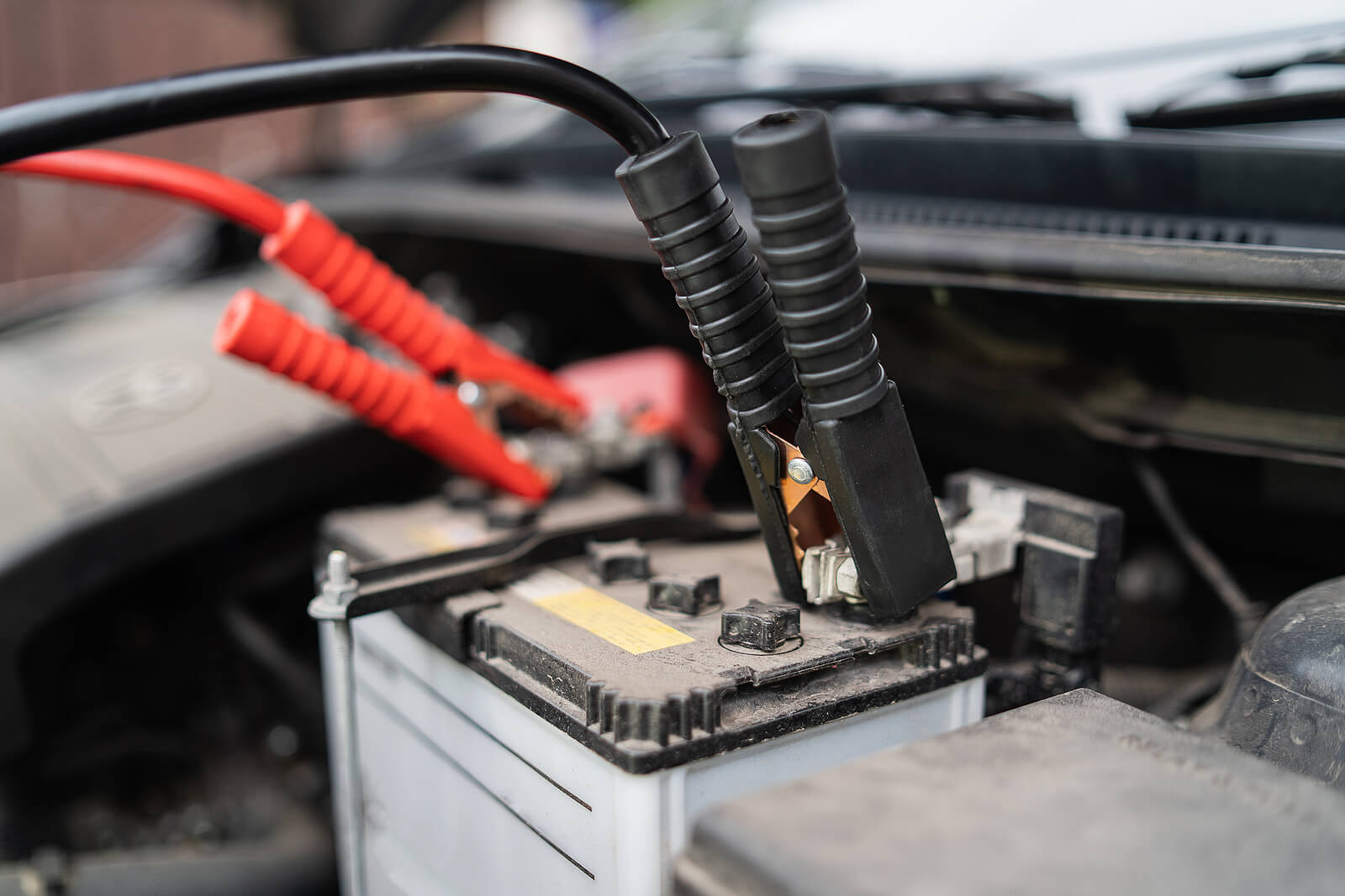 A close-up of a car battery and jump leads under a car bonnet.