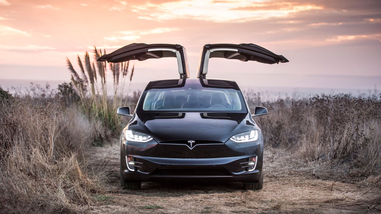 Photograph of a Tesla model X near the beach, sun is setting, doors are open.