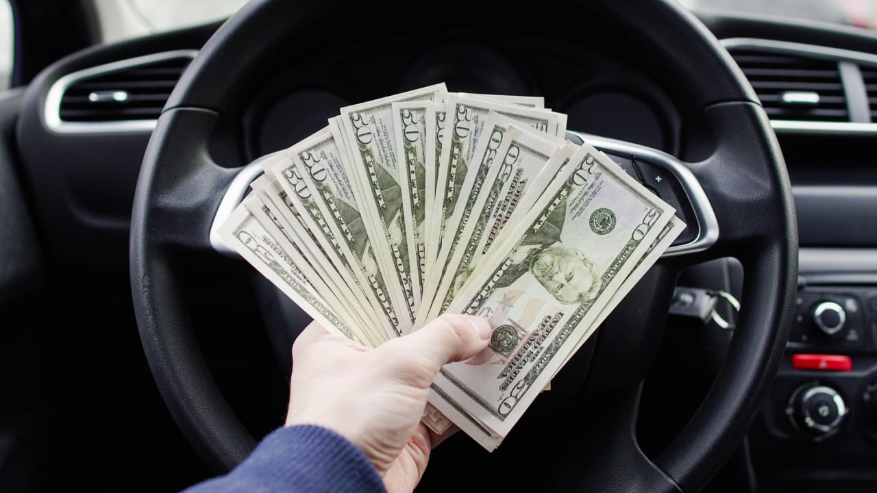 A dollar bills in the male hand on the background of the steering wheel in the car. Concept of earnings or bribes, insurance or credit, investing or selling a car. electric car battery replacement cost.
